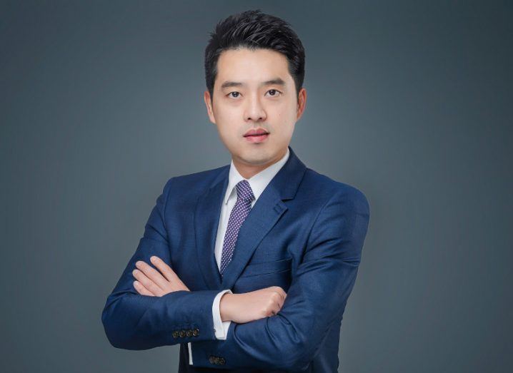 Jijay Shen, CEO of Huawei, wearing a blue suit with arms crossed, standing against a grey background.