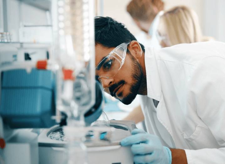 A young man in a lab looking at a piece of equipment. He is wearing goggles and a white coat.