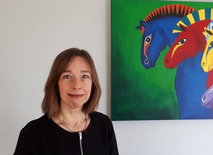 Sandra Hurley standing and smiling beside a multicoloured art piece featuring horses.