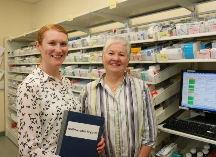 Two women stand in a pharmacy storeroom with fully stocked shelves of treatments. They are standing in front of a desktop computer and one woman holds a folder that reads ‘antimicrobial register’.
