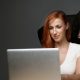 Red-headed white woman types on laptop while menacing figure wearing balaclava looks over her shoulder.