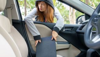 A young woman wearing a hoodie taking a laptop from the front seat of a car.